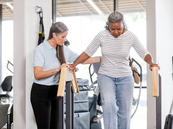 A woman helps another woman walk with parallel bars in a Medbridge rehab setting.
