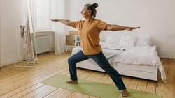 A person doing yoga in warrior pose on a green mat in a bright bedroom.