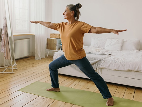 A person doing yoga in warrior pose on a green mat in a bright bedroom.
