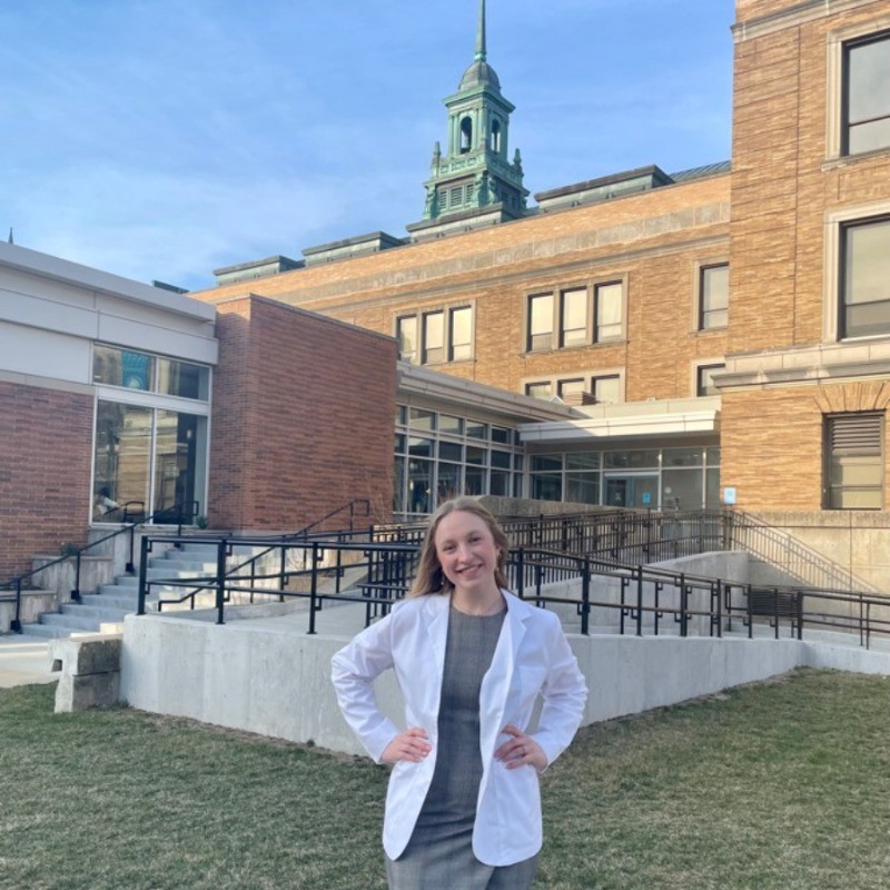 A woman in a white coat smiles outside the medbridge building with a tall spire.