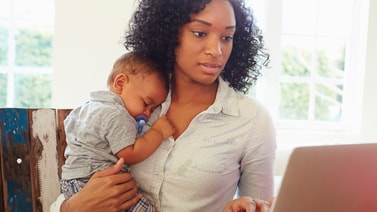 Woman with a sleeping baby works on a laptop, viewing medbridge patient care software.