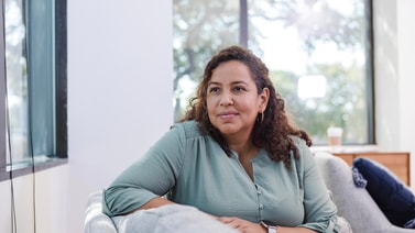 A woman with curly hair smiles gently on a bright sofa, reflecting the comfort of medbridge digital healthcare at home.