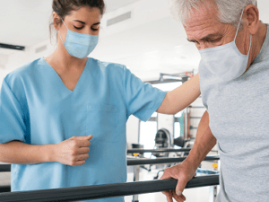 A healthcare worker helps an elderly man with balance exercises using parallel bars, while two others observe in the background; all are masked at a medbridge facility.