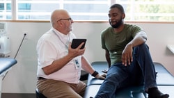 A doctor and patient converse on an exam table, with the doctor holding a tablet.