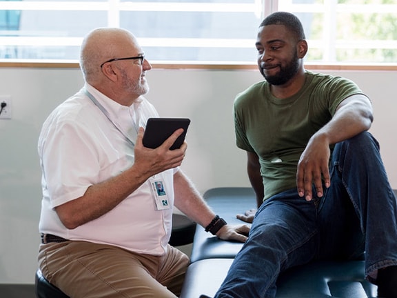 A doctor and patient converse on an exam table, with the doctor holding a tablet.