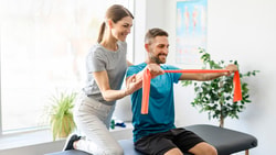 A woman assists a man with stretching using a resistance band on a black table at medbridge.