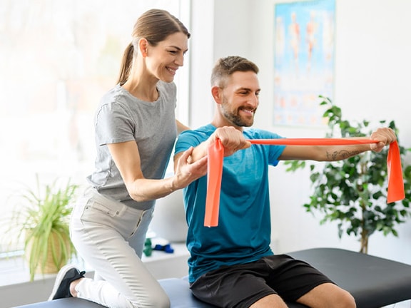 A woman assists a man with stretching using a resistance band on a black table at medbridge.