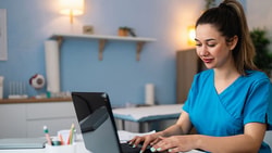 A Medbridge user in scrubs types on a laptop at a medical desk.