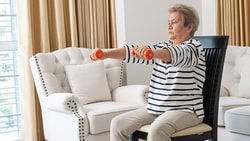 Older adult seated in a living room performing a seated dumbbell front raise exercise with small dumbbells.