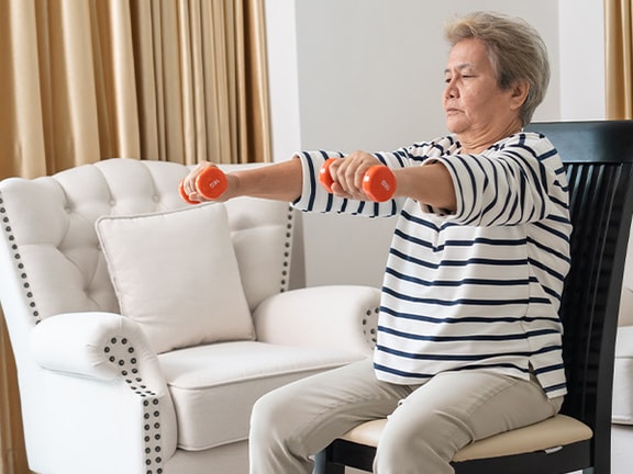 Older adult seated in a living room performing a seated dumbbell front raise exercise with small dumbbells.