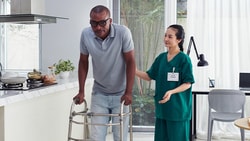 A Medbridge healthcare worker in green assists a man with a walker in a bright kitchen.