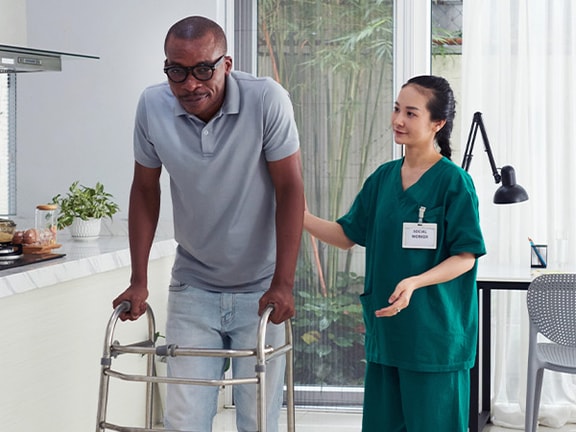 A Medbridge healthcare worker in green assists a man with a walker in a bright kitchen.