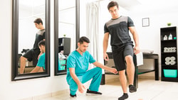 A physical therapist in blue scrubs helps a man balance on one leg in a therapy room, for medbridge.