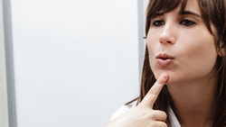 A person in an indoor setting forms an 'o' shape with their lips while touching their chin, facing another.