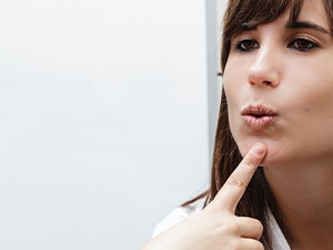 A person in an indoor setting forms an 'o' shape with their lips while touching their chin, facing another.