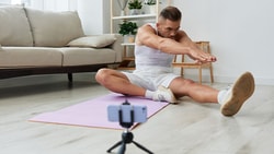 Individual doing yoga on a mat while recording with a smartphone on a tripod at home.
