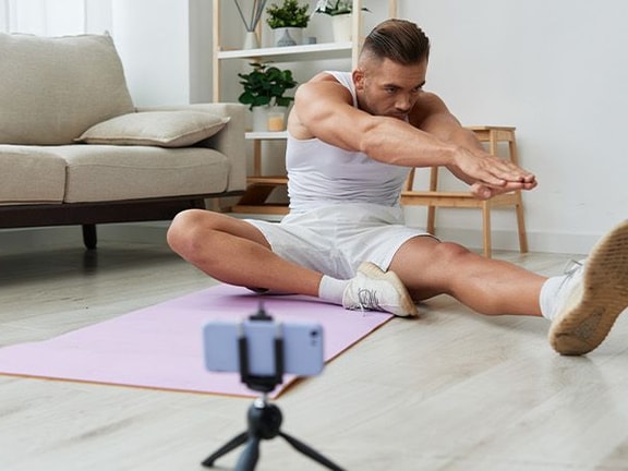 Individual doing yoga on a mat while recording with a smartphone on a tripod at home.