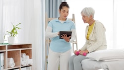 A healthcare professional in a clinic shows an older woman holding pink dumbbells a tablet from medbridge.