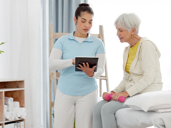 A healthcare professional in a clinic shows an older woman holding pink dumbbells a tablet from medbridge.