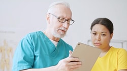 A Medbridge healthcare worker in blue scrubs shows a tablet to a concerned person in yellow.
