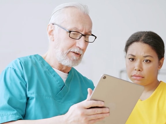 A Medbridge healthcare worker in blue scrubs shows a tablet to a concerned person in yellow.