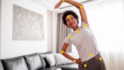 Individual stretching with motion-tracking overlay by a black sofa, reaching one arm overhead; Medbridge software visible.