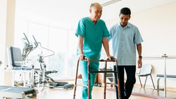 A healthcare professional helps an elderly man using a walker in a medbridge facility.