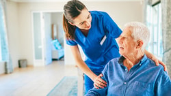 A healthcare worker in blue scrubs talks to an elderly man in a bright room.
