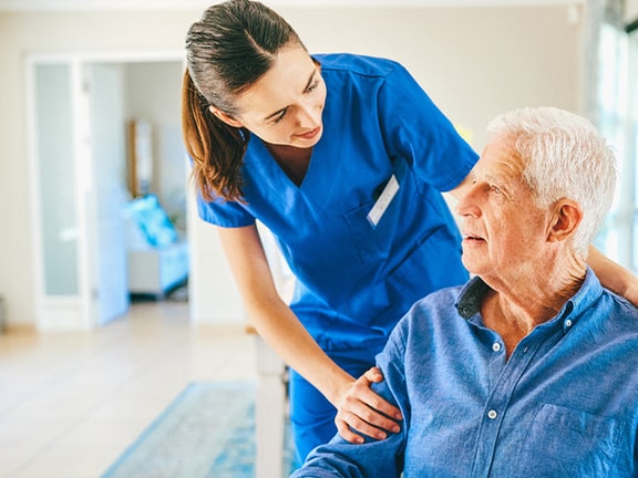 A healthcare worker in blue scrubs talks to an elderly man in a bright room.
