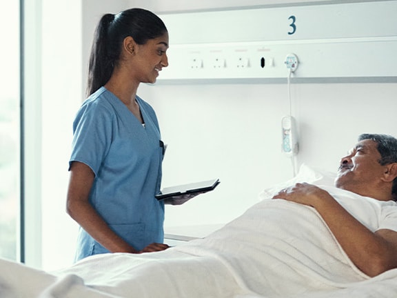 A nurse in blue scrubs holds a tablet next to a patient in a hospital bed by a window, illustrating Medbridge technology use.