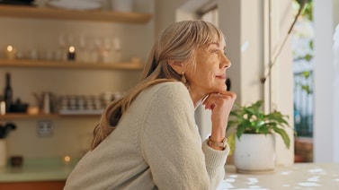 Older woman with long gray hair looks thoughtfully out a window, with medbridge content open on her device.