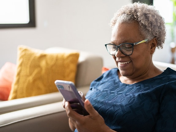 An older person with glasses smiles at their smartphone, seated on a sofa in a bright room with colorful pillows.