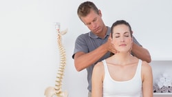 A man adjusts a woman's neck in a white room with a spine model and shelves.