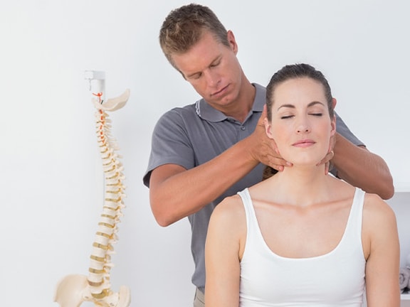 A man adjusts a woman's neck in a white room with a spine model and shelves.