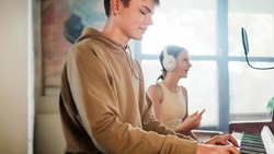 A person plays the piano as another listens on headphones holding a phone, with a microphone set up nearby.