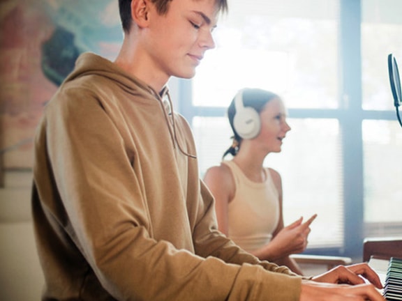 A person plays the piano as another listens on headphones holding a phone, with a microphone set up nearby.