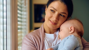 A woman holds a sleeping baby by a window, reflecting on digital healthcare and medbridge's support for families.