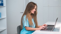 A woman in a blue uniform types on a laptop at her desk; Medbridge files are visible on the shelves behind her.