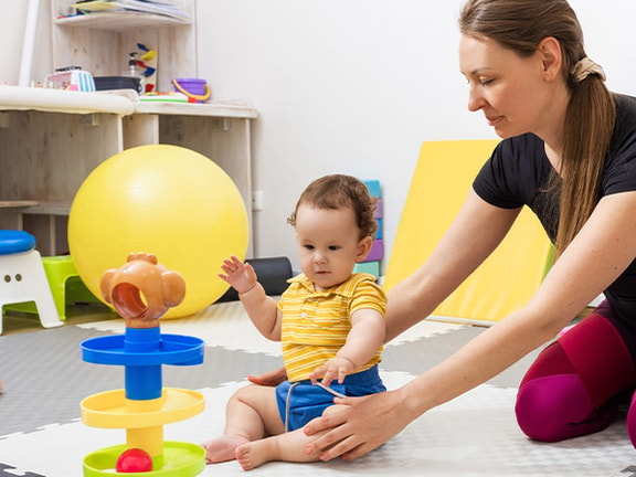 A woman and a toddler play in a colorful playroom with balls, mats, and foam blocks.