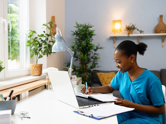 A woman in scrubs writes in a notebook at a desk with a laptop, clipboard, plants, and home decor.