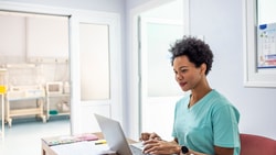 A healthcare professional in scrubs uses a laptop near medical equipment and open doors in a well-lit room.