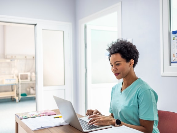 A healthcare professional in scrubs uses a laptop near medical equipment and open doors in a well-lit room.