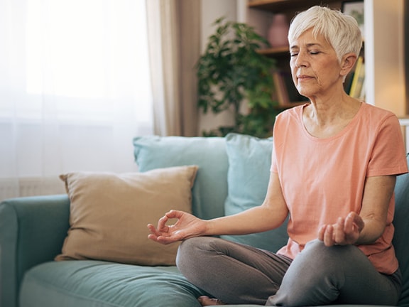 An older woman meditates on a couch with eyes closed in a bright living room.