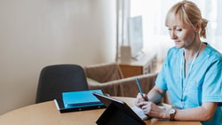 A person in blue scrubs writes on a clipboard, referencing a tablet, with a blue folder nearby.