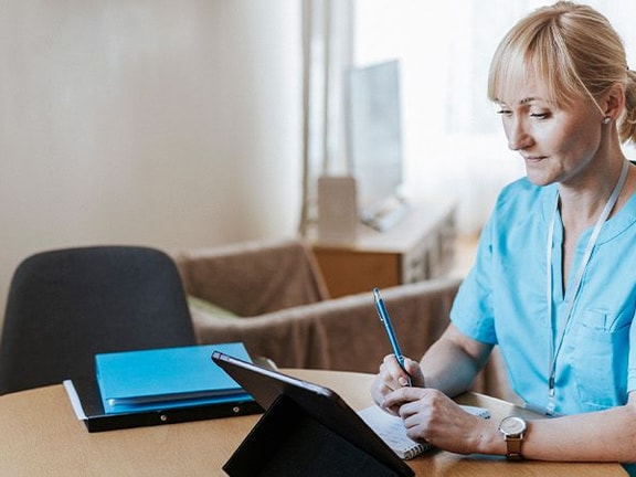 A person in blue scrubs writes on a clipboard, referencing a tablet, with a blue folder nearby.