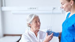 Nurse in blue scrubs standing by an elderly woman holding a water glass at medbridge.