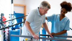 A nurse aids an elderly man using parallel bars in a well-lit room for physical therapy during a medbridge session.
