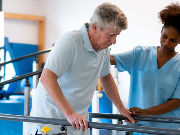 A nurse aids an elderly man using parallel bars in a well-lit room for physical therapy during a medbridge session.