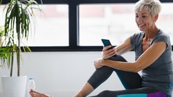 A person in workout clothes sits smiling with a smartphone near a water bottle and plant; large windows are behind them.