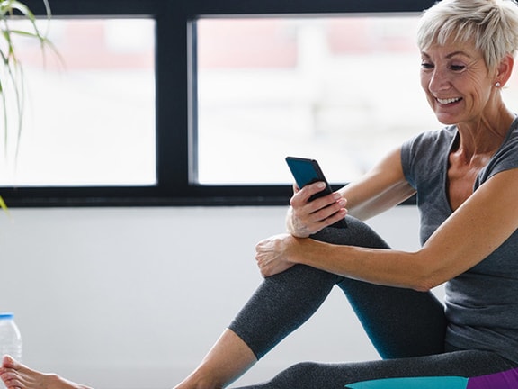 A person in workout clothes sits smiling with a smartphone near a water bottle and plant; large windows are behind them.